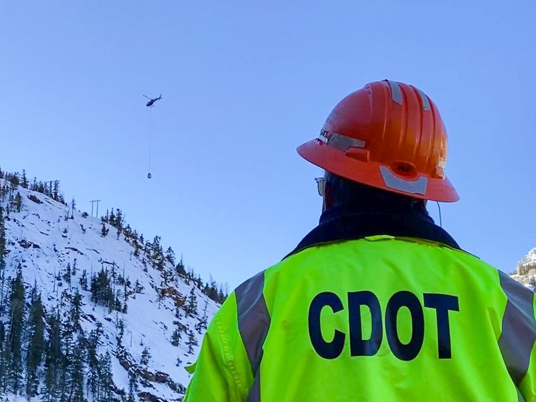 A member of CDOT's specially trained avalanche crew observes avalanche mitigation equipment being transported into place by a helicopter.